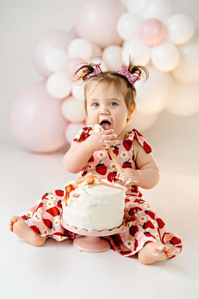 A baby with blue eyes and pigtails, wearing a strawberry-patterned outfit, sits on the floor enjoying a white frosted cake with her hands. Pink and white balloons are blurred in the background.