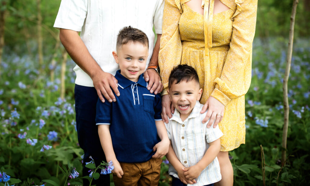 A family photo shows two young boys standing in front of two adults in a field of green plants with purple flowers. The adults’ faces are out of frame; one wears white, the other a yellow dress. The boys are smiling.