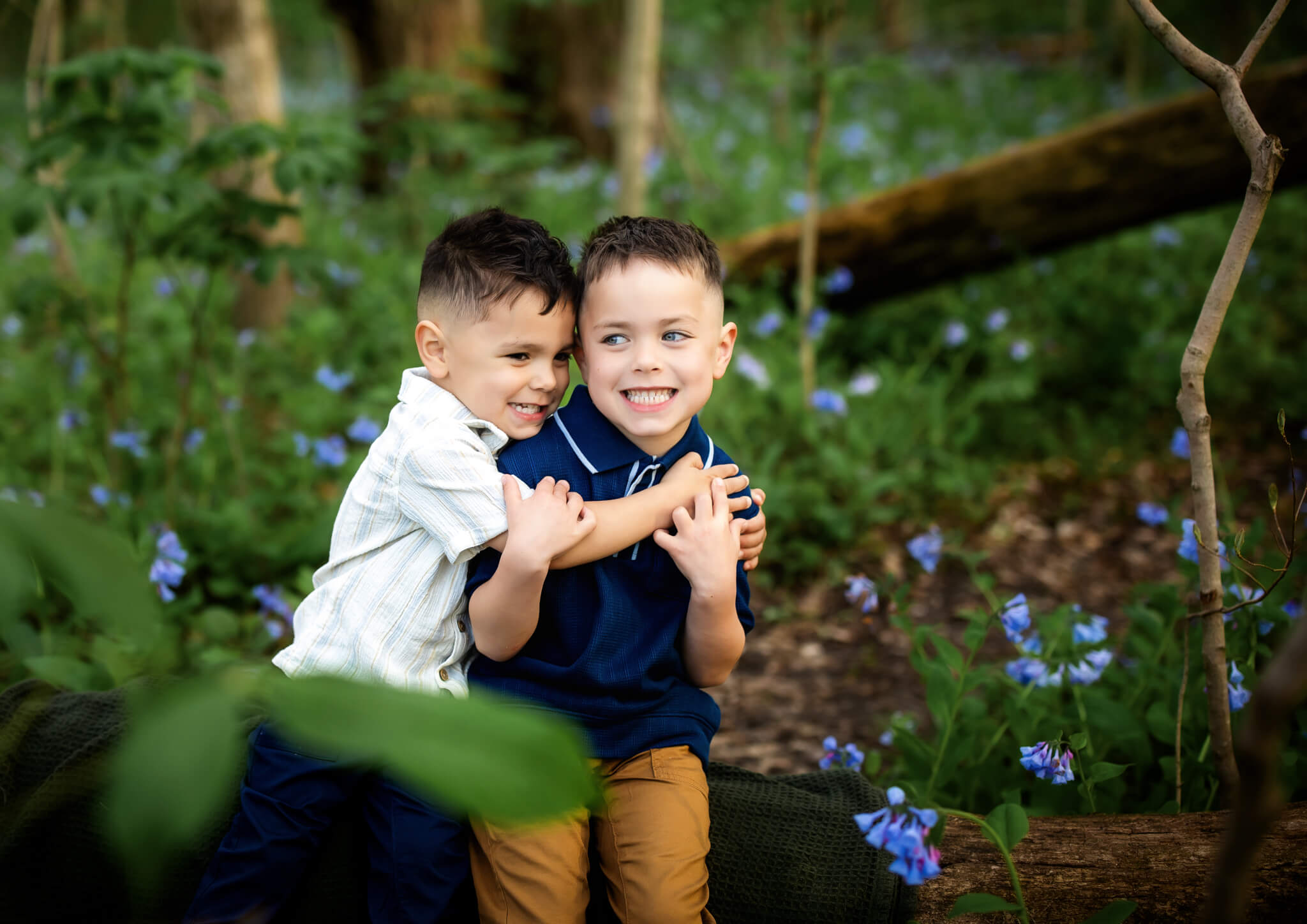 Two young boys sit close together on a log in a forest, smiling and hugging each other at bloomington normal parks. They are surrounded by green foliage and small blue flowers, enjoying a playful moment outdoors.
