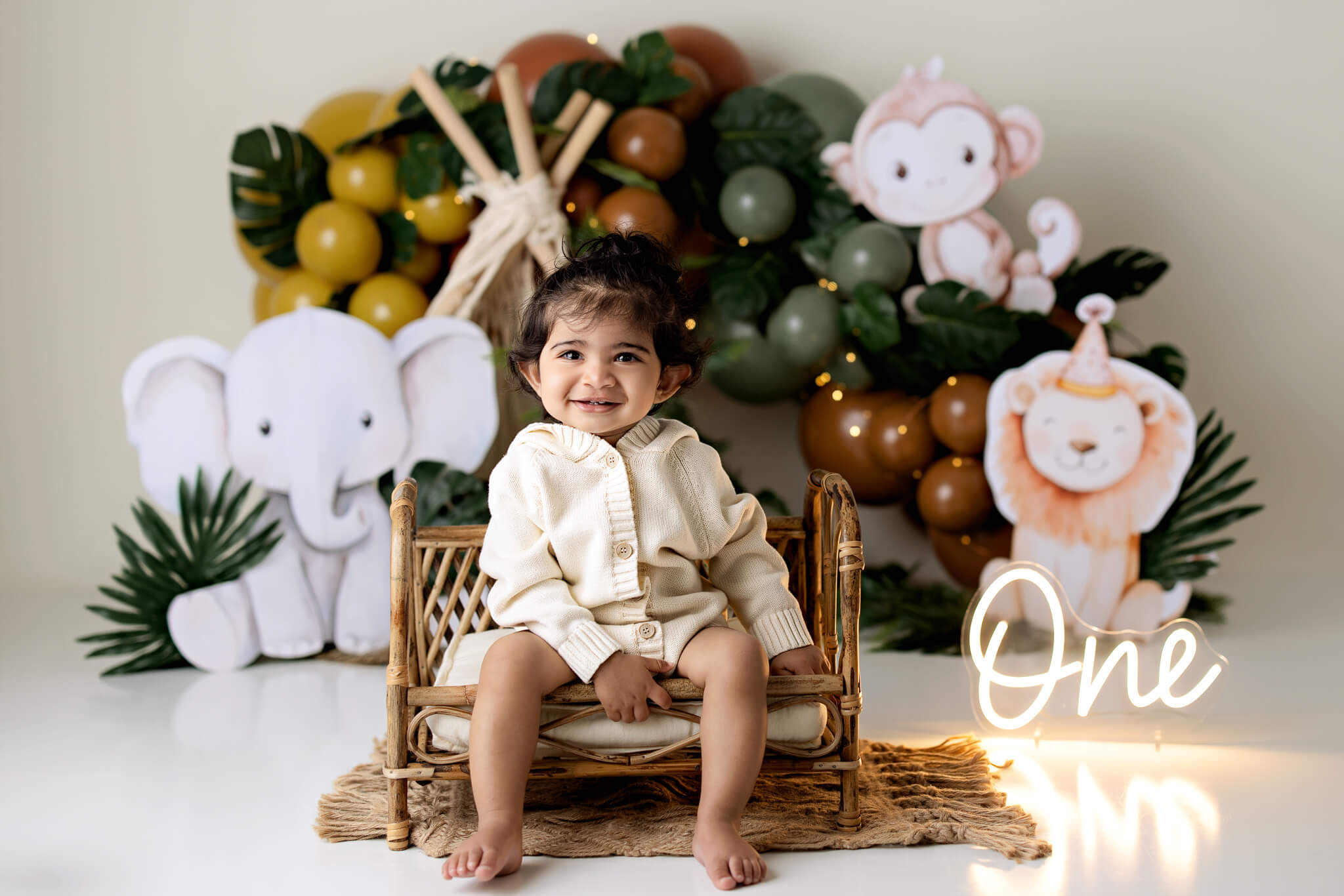 A smiling toddler sits on a wicker bench in front of jungle-themed decorations, captured by a Bloomington family photographer. Cartoon elephants, monkeys, and lions surround the glowing word "One," setting the scene for a joyful first birthday celebration.