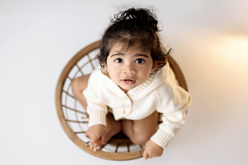 A young child with dark hair and expressive eyes, wearing a white sweater, sits in a round wooden basket and looks up toward the camera against a light background—a charming moment captured by a skilled Bloomington family photographer.