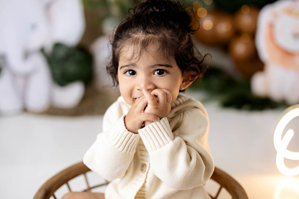 A young child with dark, curly hair and big brown eyes sits in a chair wearing a cream-colored sweater, holding their hands near their mouth and looking at the camera. Plush animals are blurred in the background—captured by a Bloomington family photographer.