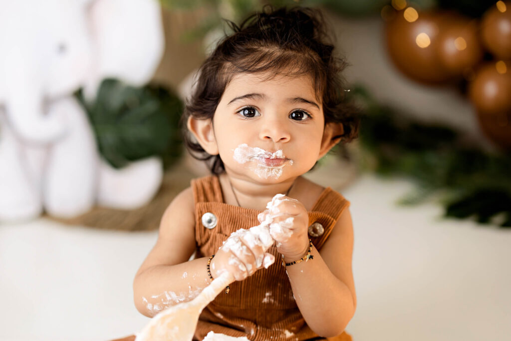 A young child in brown overalls sits indoors, holding a wooden spoon with white frosting smeared on their face and hands. Captured by a talented children’s photographer, plush decorations and green leaves create a charming blurred background.