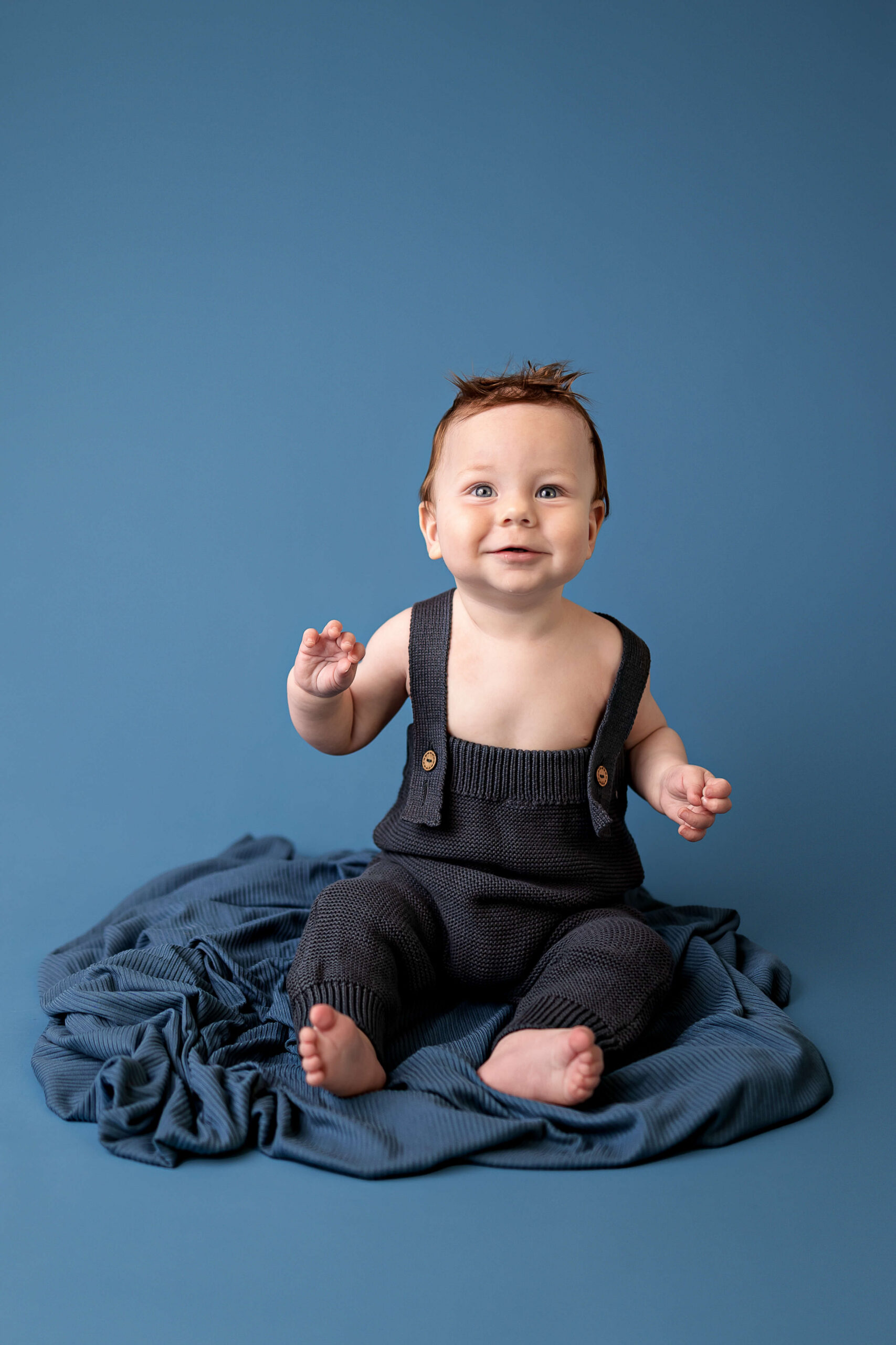 A smiling baby in blue overalls sits on a blanket in a studio