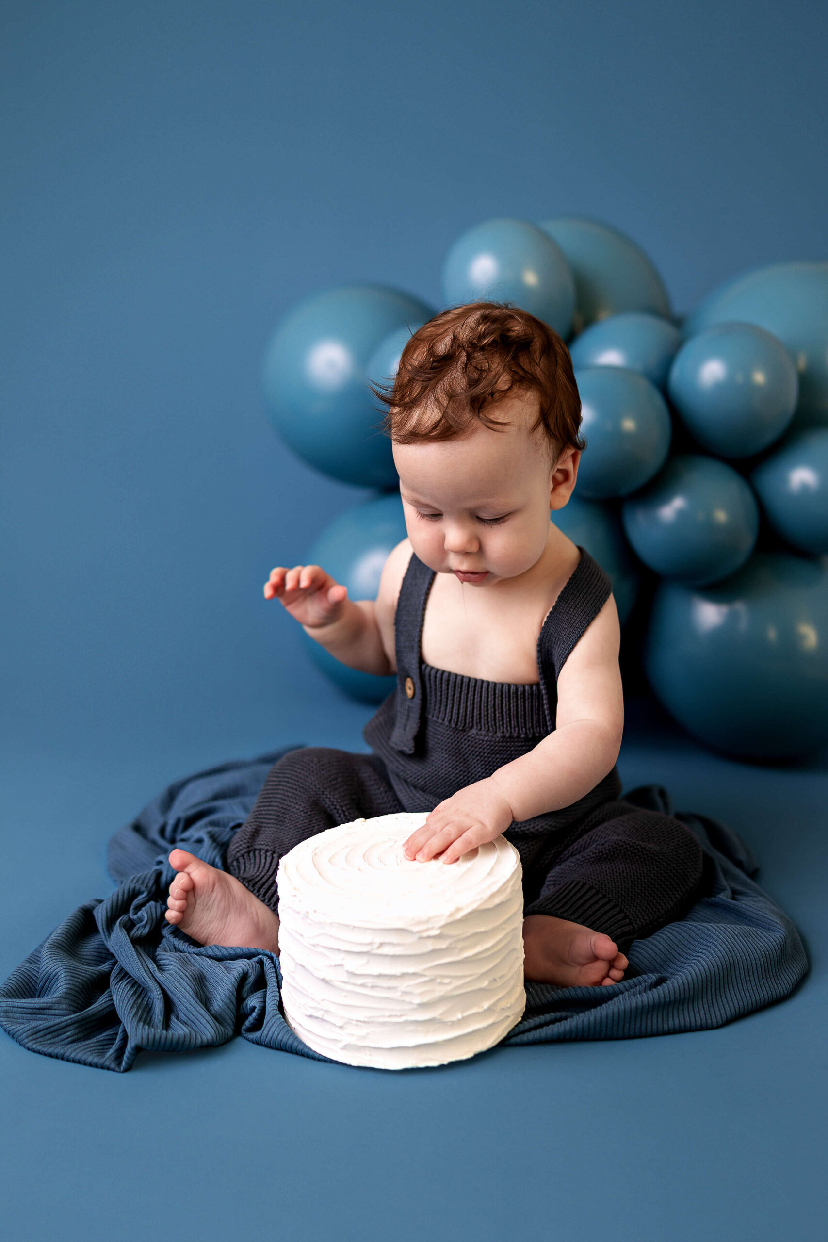 A baby blue in blue overalls drools over a white cake with his hand on it for his birthday