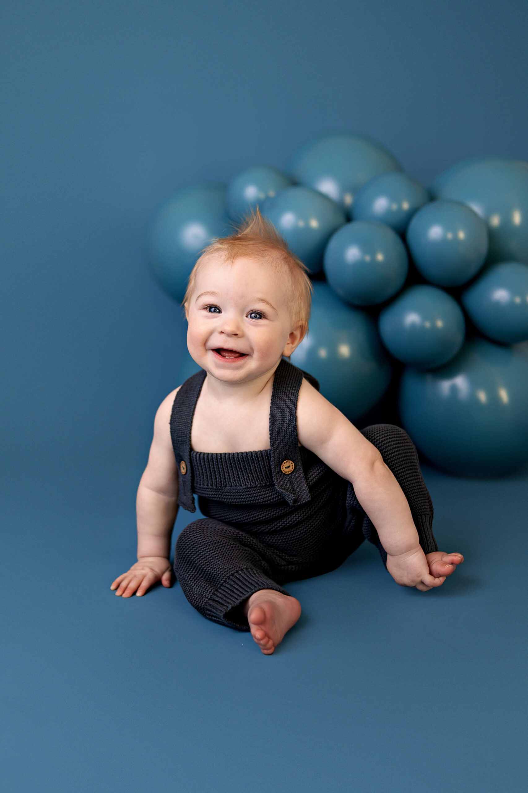 A happy baby boy in blue knit overalls sits holding his foot in a studio before visiting the Normal Public Library