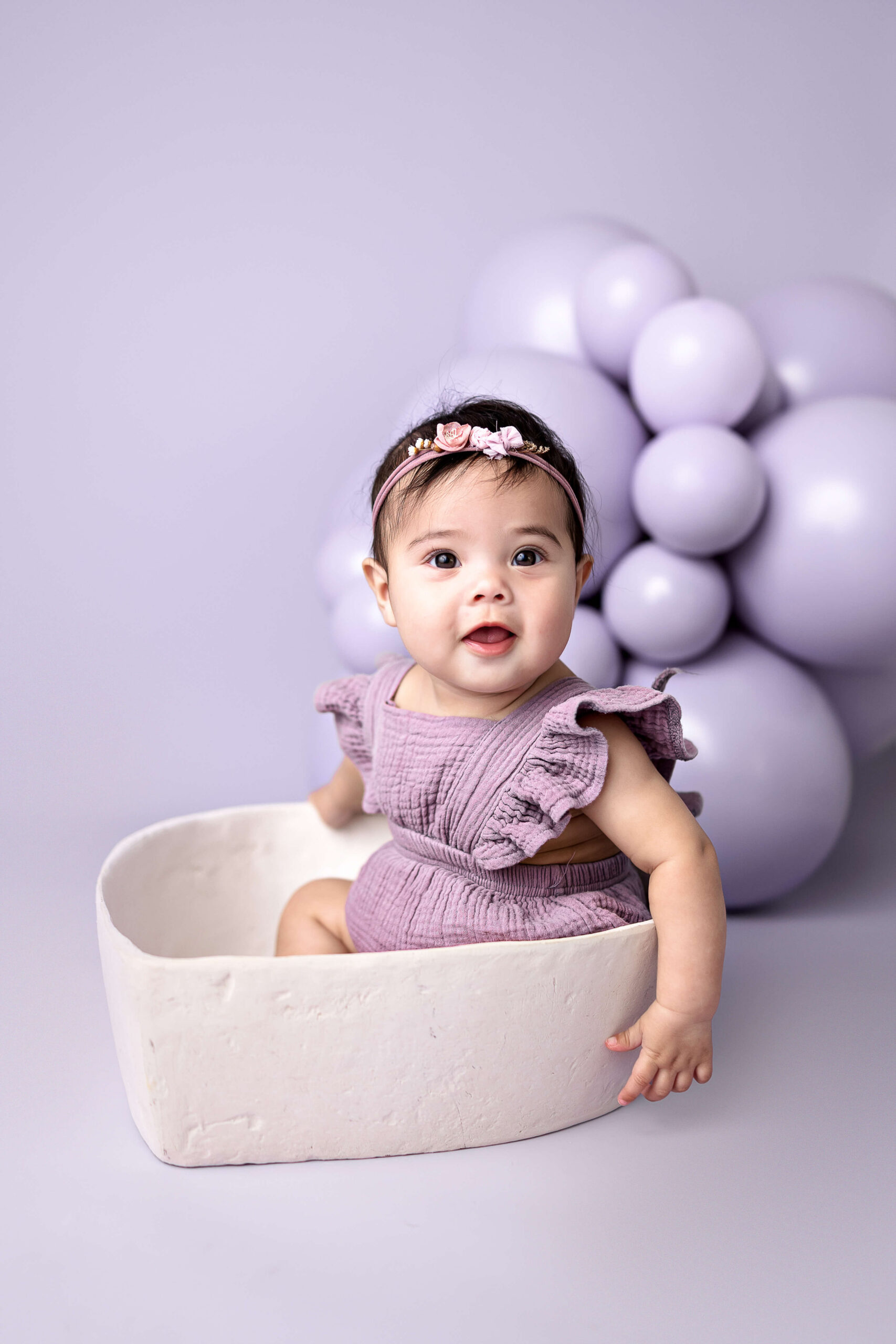 A happy baby sits in a bowl in a purple dress in front of matching balloons with a flower headband before visiting Normal Public Library