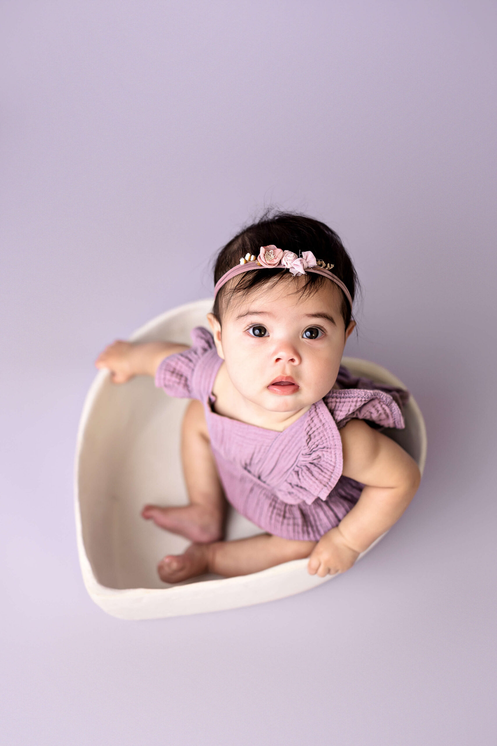 A baby looks up from sitting in a heart shaped bowl in a purple dress in a studio
