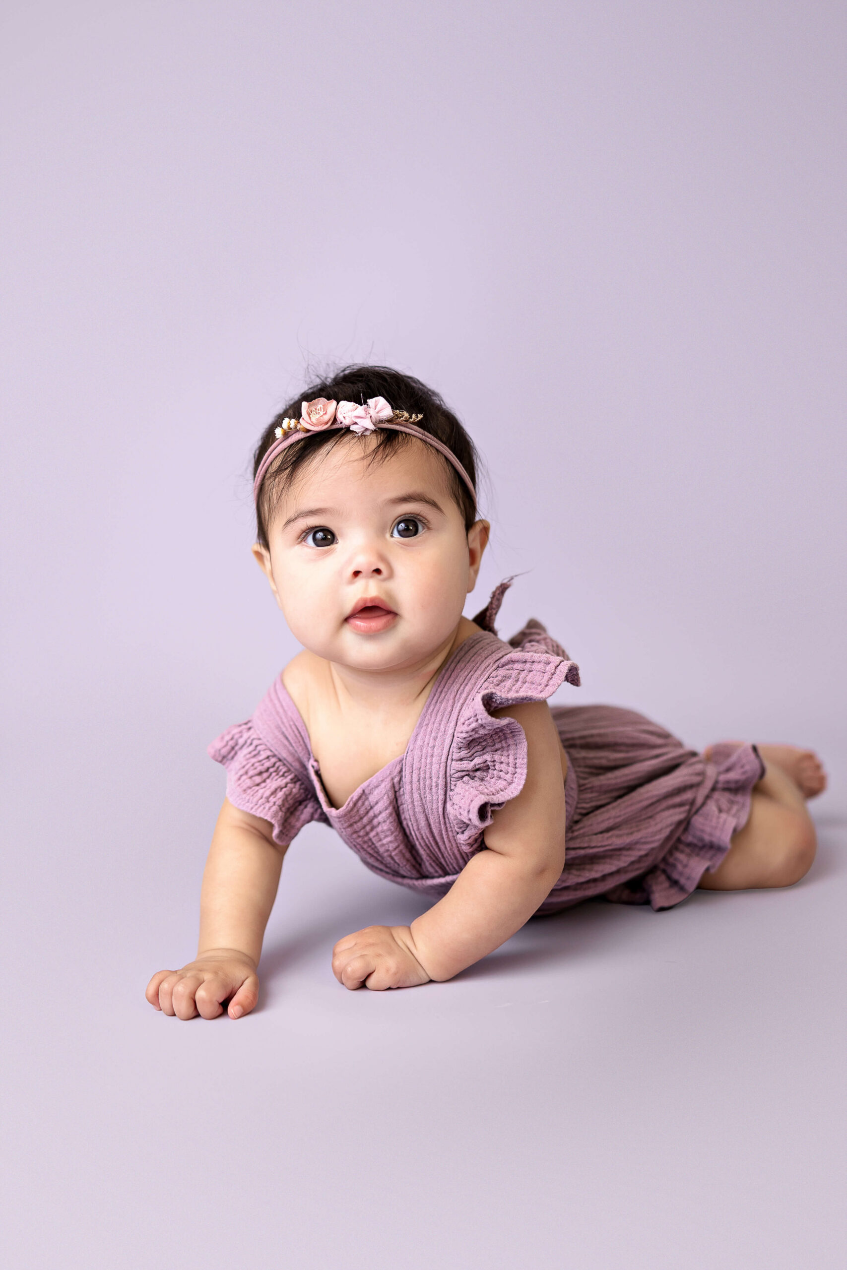A baby girl in a purple dress crawls on the floor of a matching studio with a flower headband before visiting Normal Public Library